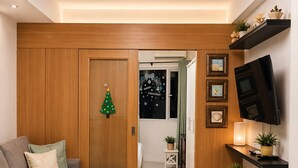 A modern living room with a sectional sofa, a wooden door with a Christmas decoration, a flat-screen TV mounted on the wall, and a small table with a candle. - Luxe 1-bedroom Condo Front of Mall of Asia With City View in Pasay (Pasay)