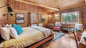 L’intérieur d’une cabane en rondins, avec un lit, une chaise, une table et une fenêtre donnant sur la forêt.
