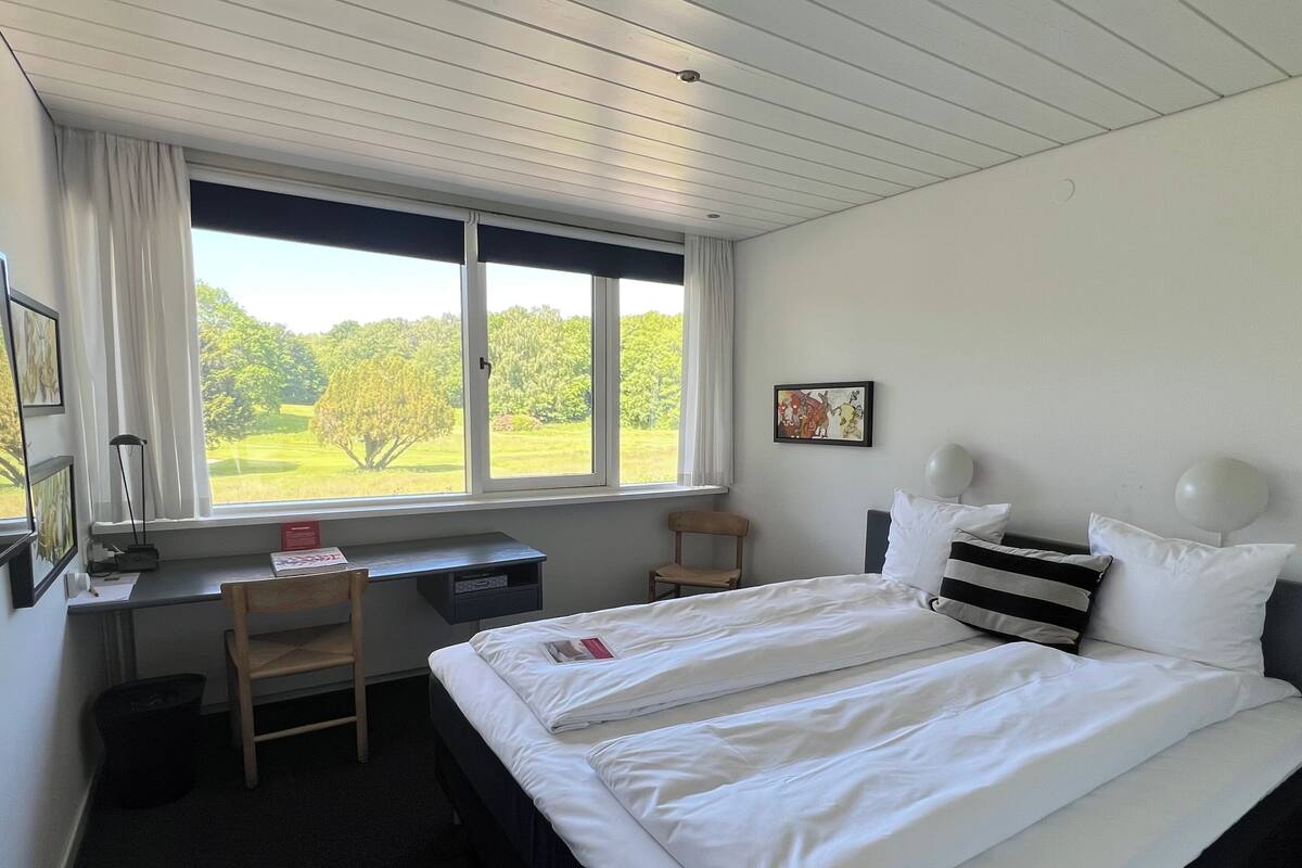 A hotel room with a bed, a desk, a chair, a window with a view of greenery, and a wall-mounted lamp.
