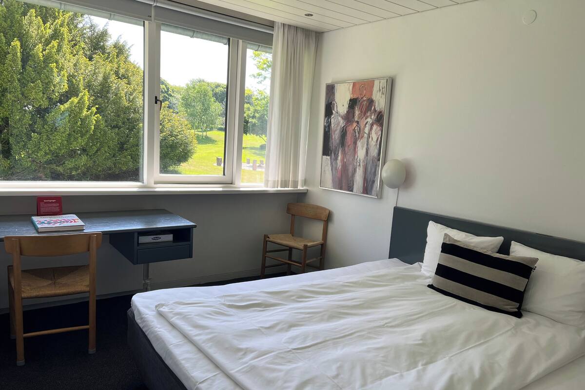 A hotel room with a bed, a desk, a chair, a window with a view of trees, and a painting on the wall.