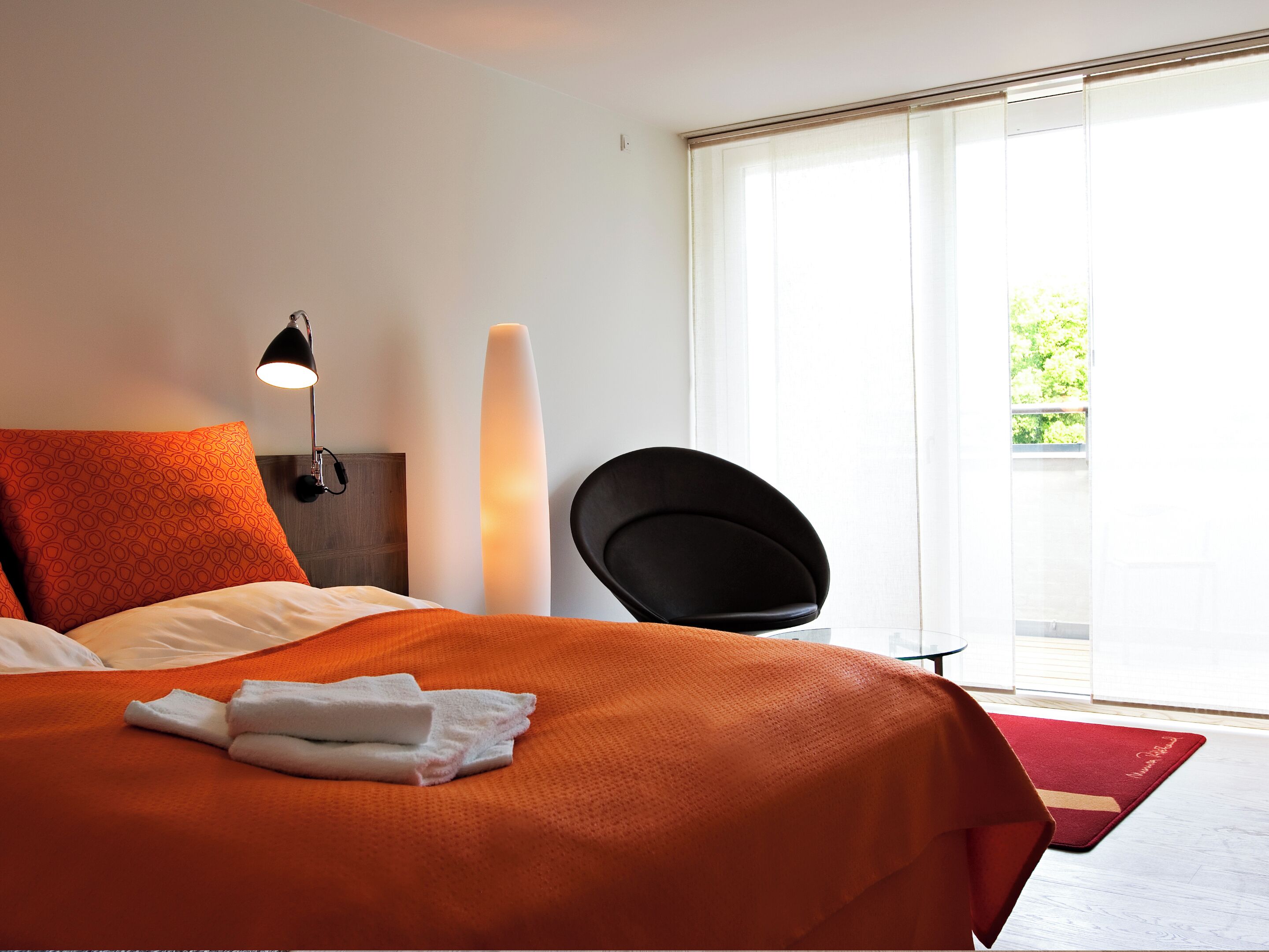 A hotel room with a bed, a standing lamp, a chair, and a view of greenery outside.