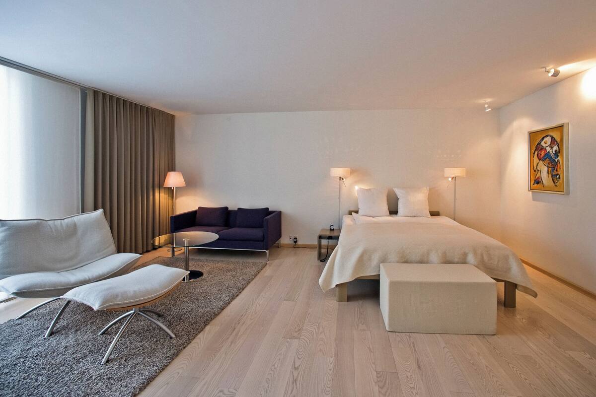 A modern hotel room with a bed, a sofa, a small table, and a lamp.