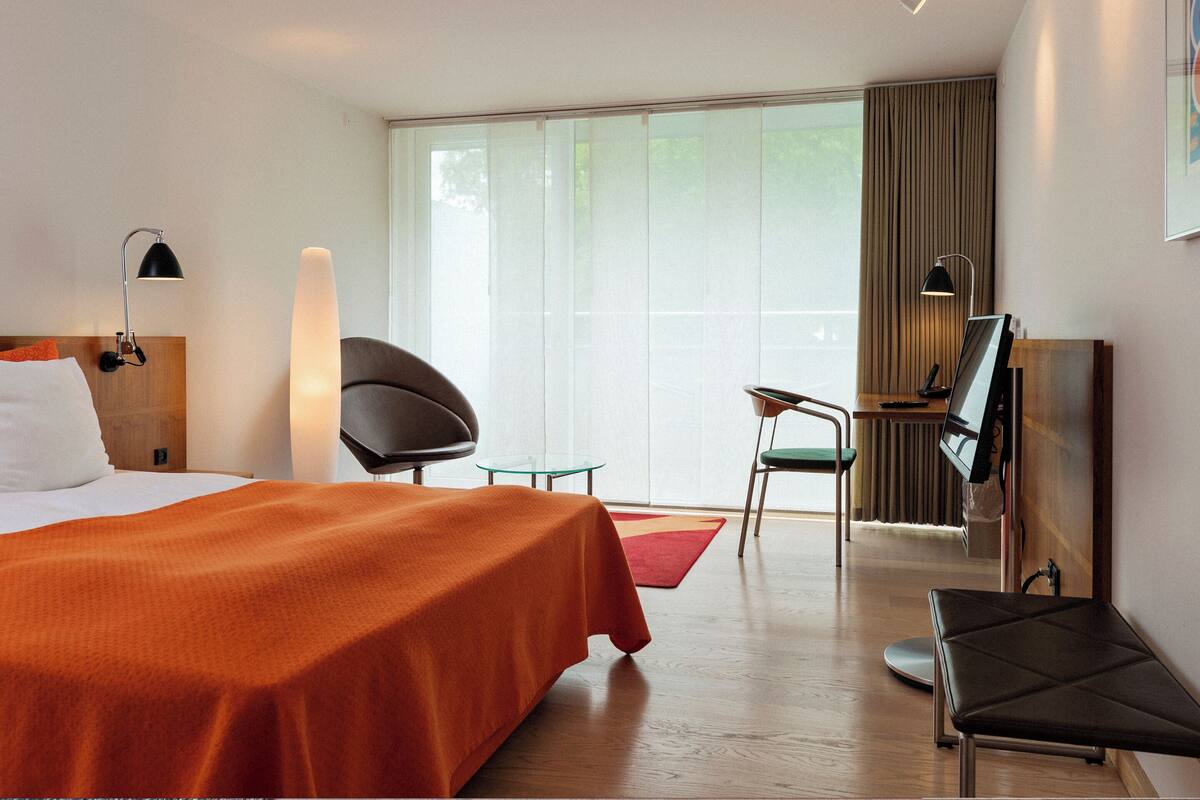 A hotel room with a bed, a chair, a desk, a TV, and a large window.