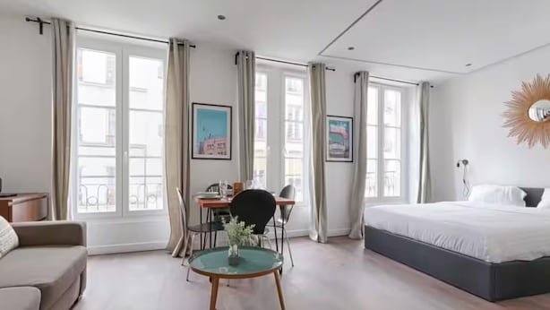 A modern hotel room with a large bed, a dining table with chairs, a small round table with a vase, and large windows with curtains. - UrbanFlat 203 + 202 - 249 Saint Denis SD1 (Paris)