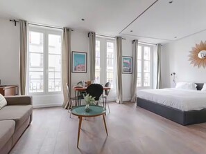 A modern hotel room with a large bed, a dining table with chairs, a small round table with a vase, and large windows with curtains. - UrbanFlat 203 + 202 - 249 Saint Denis SD1 (Paris)