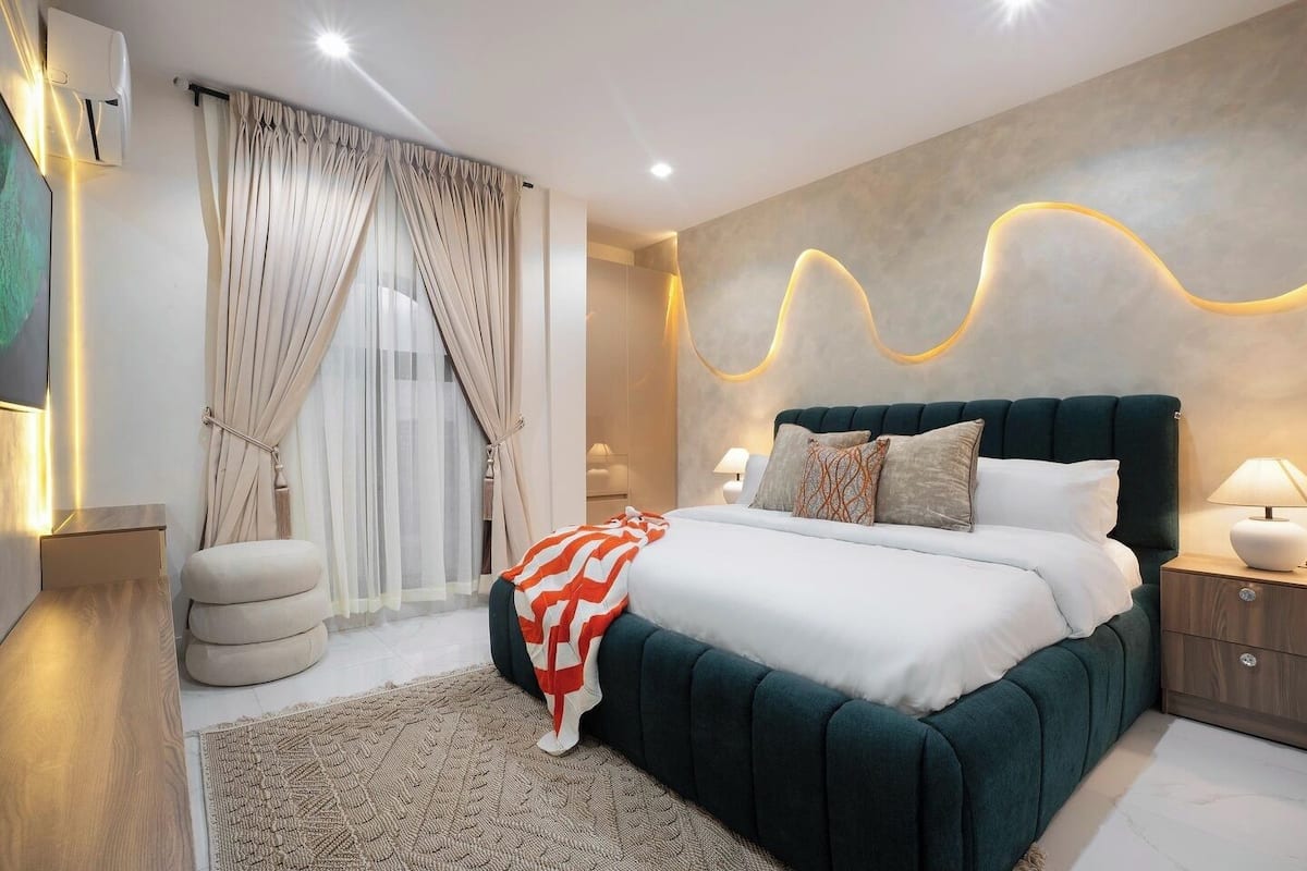 Quarto moderno com uma cama grande, mesa de cabeceira, luminária na parede e janela com cortinas.