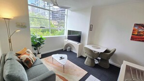 Ein modernes Wohnzimmer mit einer Couch, einem Couchtisch, einem Fernseher auf einem Schrank, einer Pflanze und einem Fenster mit Blick auf die Stadt.