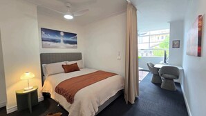 Ein Hotelzimmer mit Bett, Nachttisch, Lampe, einem Gemälde, Blick nach draußen und einem kleinen Tisch mit Stühlen.