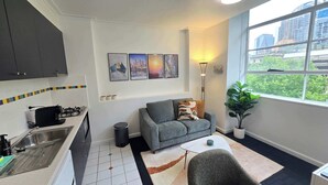 Ein kompakter Wohnbereich mit Küchenzeile, grauem Sofa, einem kleinen weißen Tisch und einem großen Fenster mit Blick auf die Stadt.