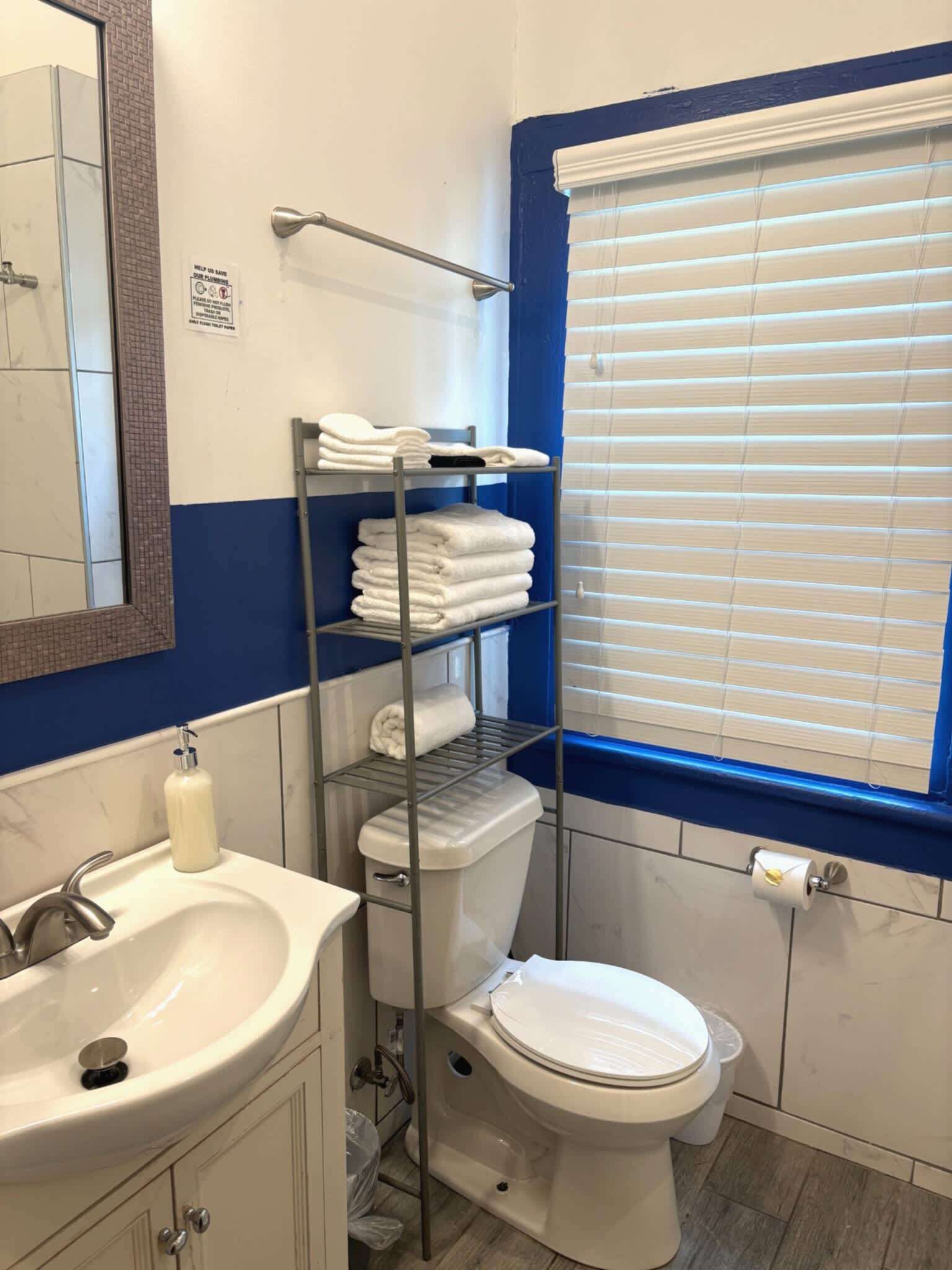 Une salle de bain comprenant des toilettes, un lavabo, un miroir, un porte-serviettes et une fenêtre avec des stores.