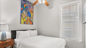 Ein Schlafzimmer mit einem Bett, einem Nachttisch, einer Lampe, einem farbenfrohen abstrakten Gemälde und einem Fenster mit Jalousien.