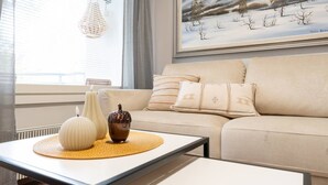 Ein Wohnzimmer mit einem beigefarbenen Sofa, dekorativen Kissen, einem weißen Tisch mit Vasen und einem Gemälde von Winterlandschaft.