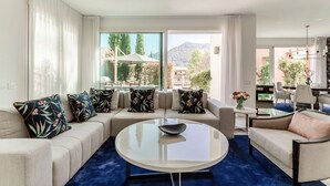Un salon moderne comprenant un ensemble de canapés, une table basse ronde et une vue sur un jardin et des montagnes, grâce à de grandes fenêtres.