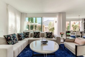 Ein modernes Wohnzimmer mit einer Sofagarnitur, einem runden Couchtisch und Blick auf einen Garten und Berge durch große Fenster.