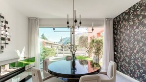 Un espace repas comprenant une table ronde, des chaises, un lustre et une vue sur les bâtiments et la verdure environnante.