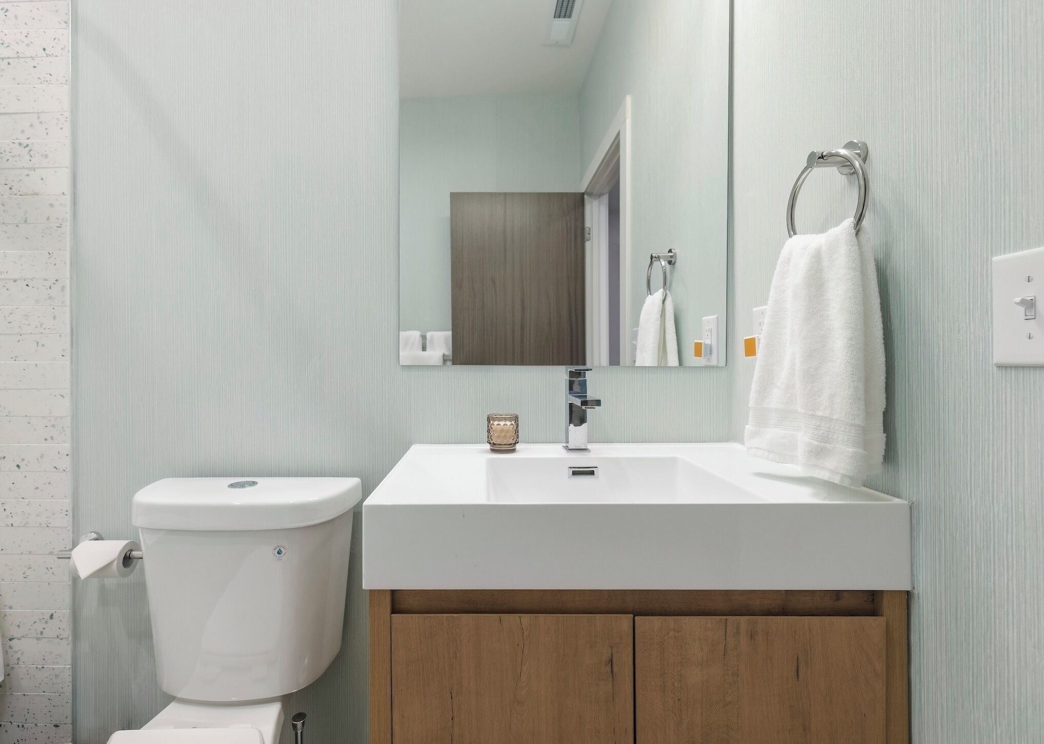 Une salle de bain avec des toilettes blanches, un lavabo surmonté d’un miroir et une serviette suspendue à un anneau en métal.