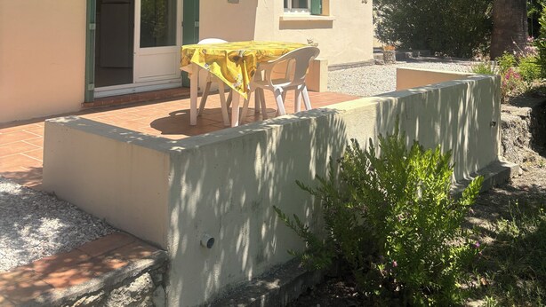 Ein Gebäude mit einer Terrasse, einem Tisch mit Stühlen und einer Tür mit grünen Fensterläden.