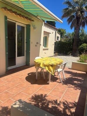Una casa con un patio, una mesa con mantel amarillo, una puerta con contraventanas verdes y una palmera.