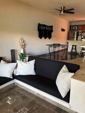 Ein Wohnzimmer mit schwarzem Sofa, einem kleinen Esstisch, einem Deckenventilator und einer Küchenzeile im Hintergrund.