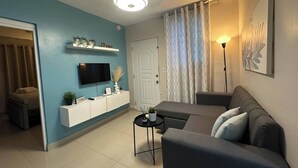 A living room with a sofa, a TV mounted on a white cabinet, a ceiling fan, a door, and a patterned curtain. - Sunset Place One Bedroom in Front of Pucmm (Santiago de los Caballeros)