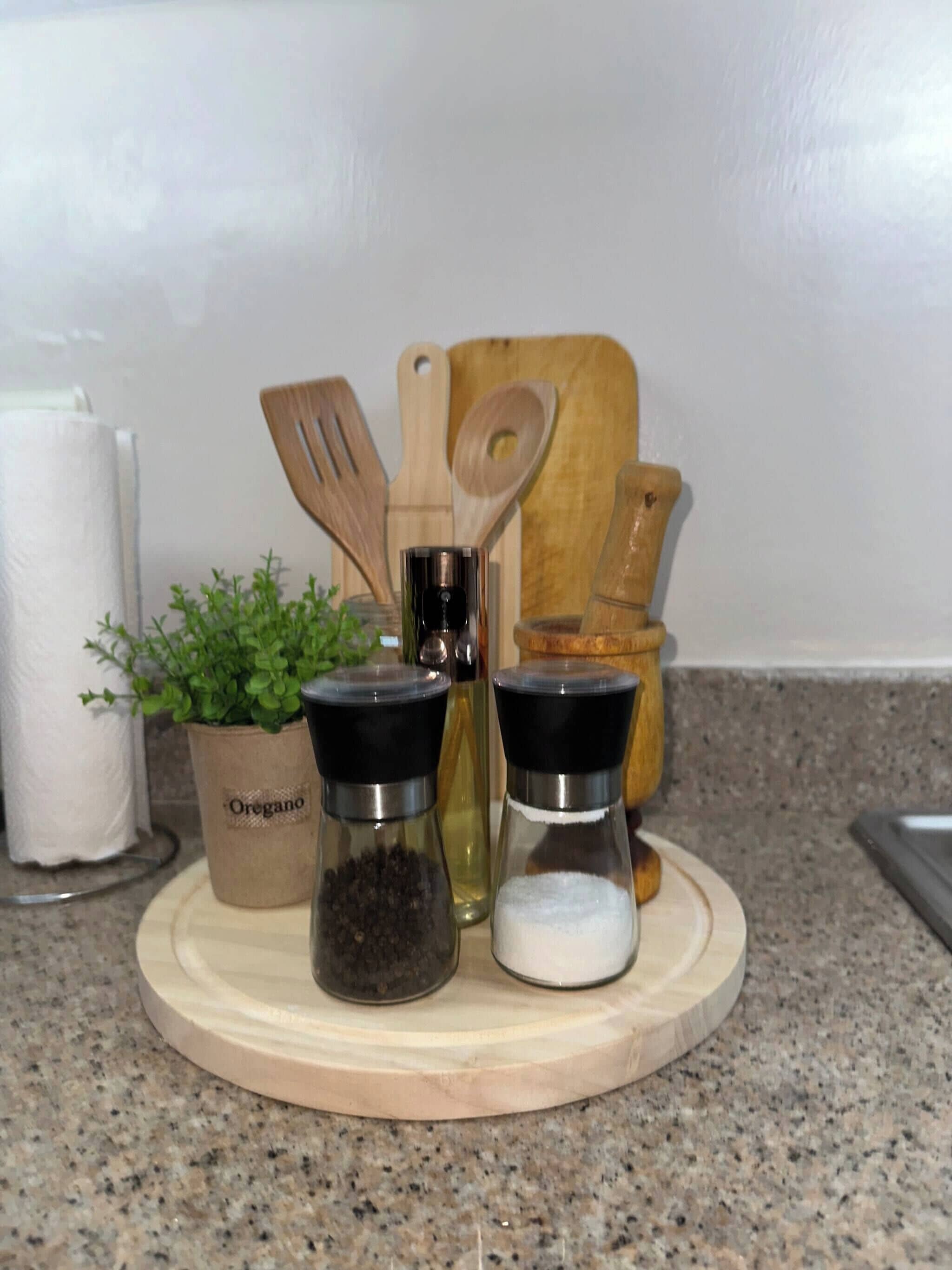 Una encimera de cocina con una tabla de cortar de madera, una planta etiquetada como 'Oregano' y dos frascos de especias.