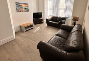 Ein Wohnzimmer mit einem braunen Ledersofa, einem Fernsehschrank, einer Bank und einem Bild an der Wand.