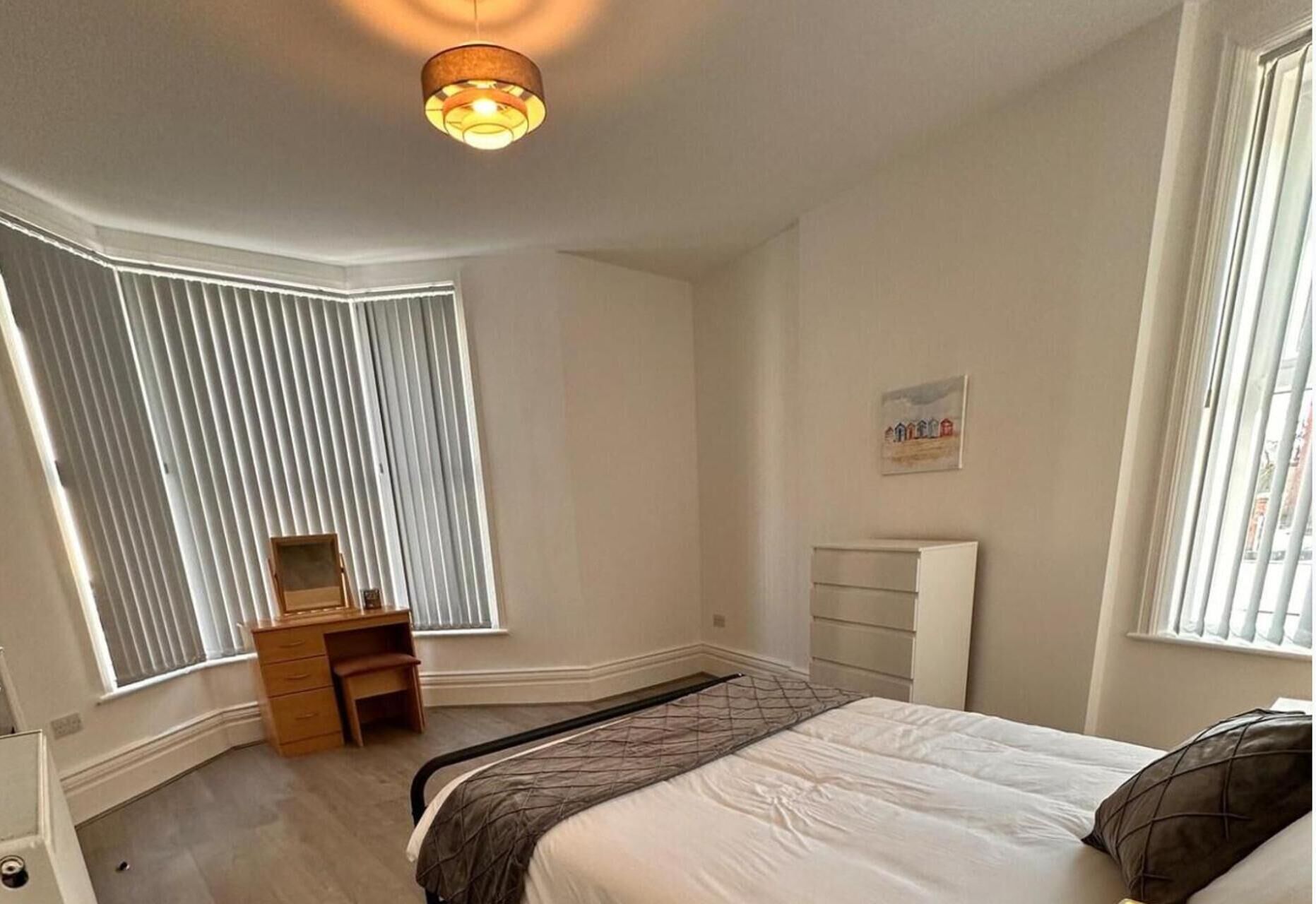 A bedroom with a bed, a dresser, a desk, a radiator, and a window with blinds.