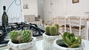 Eine Küche mit Gasherd, drei Blumentöpfen, einem Esstisch mit Stühlen und einem Kühlschrank im Hintergrund.