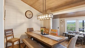 Un comedor con una mesa de madera, sillas, un banco, un reloj y vistas a la vegetación por la ventana.