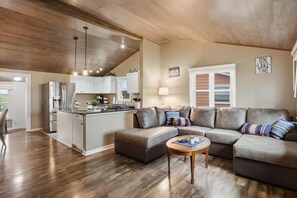 Una cocina moderna con una isla central, una zona de comedor con un sofá gris y una mesa de centro, y una sala de estar con un sofá modular gris.
