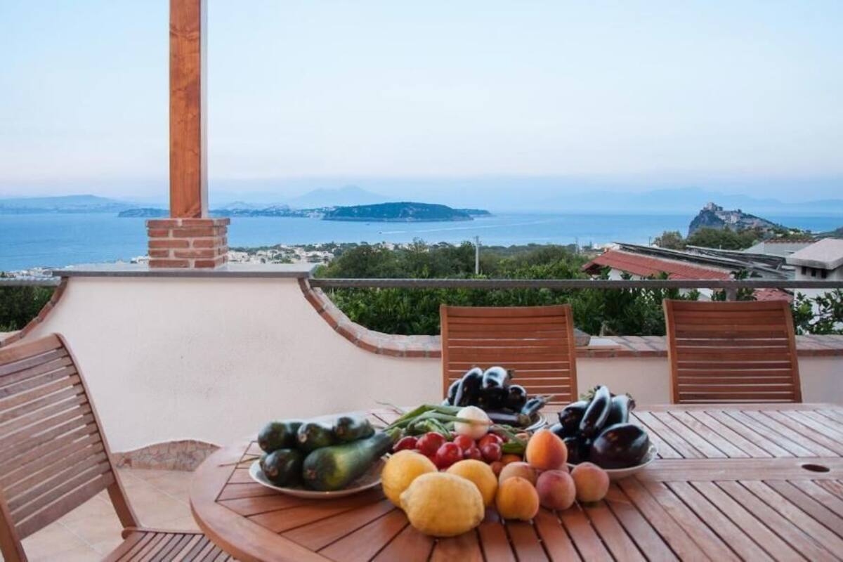 A table with fruits and vegetables, two wooden chairs, and a view of the sea and distant land.