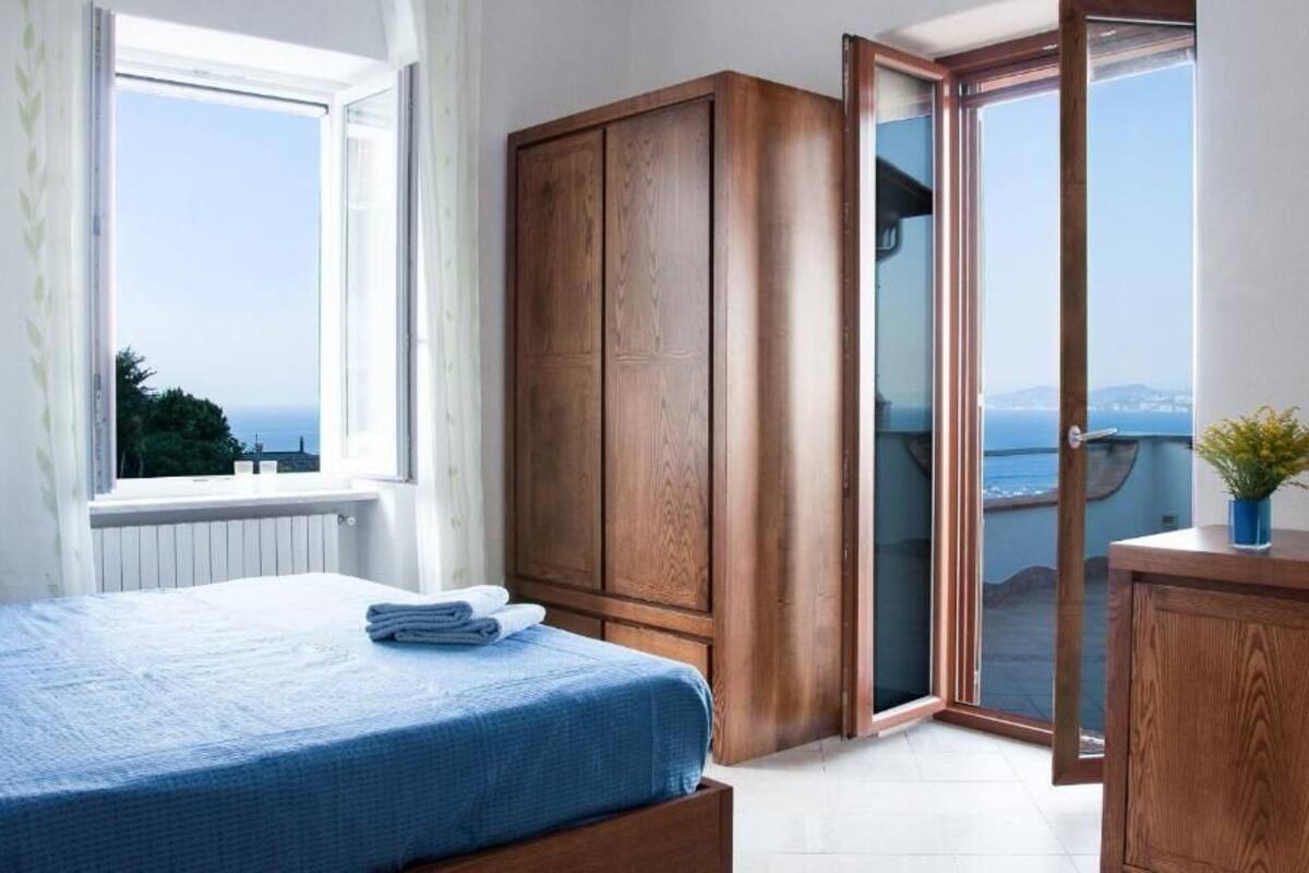 A bedroom with a bed, a wooden wardrobe, a desk, and a view through a glass door.