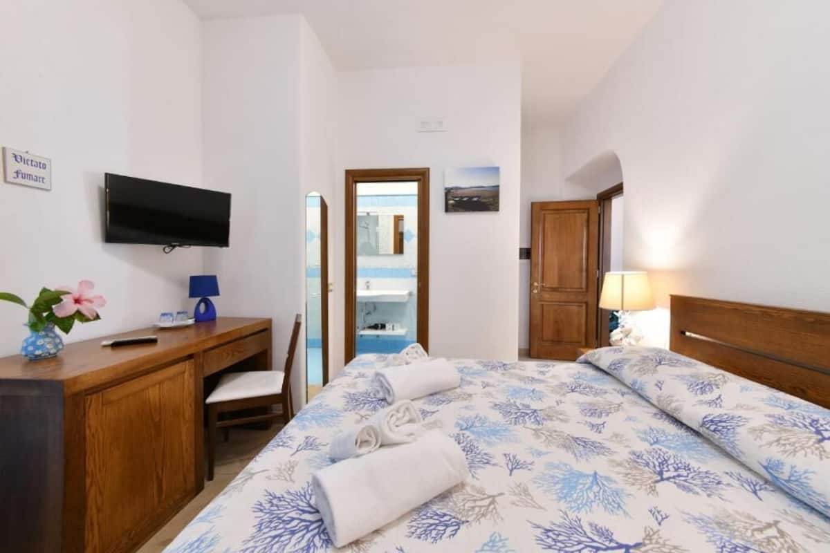 A hotel room with a bed, a desk, a television, a mirror, a door, and a bathroom visible through an open door.