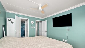Une chambre à coucher comprenant un lit, un ventilateur de plafond, un téléviseur et une porte donnant sur une salle de bain.