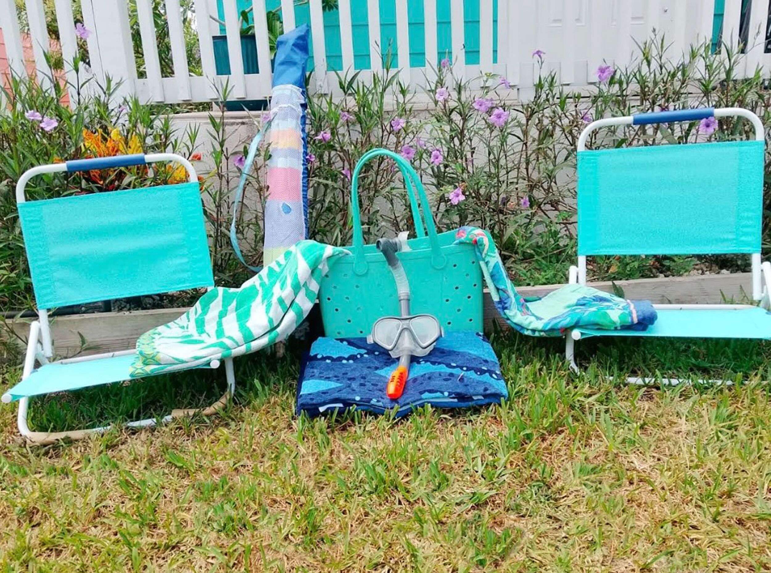 To udendørs stole med turkise hynder, en blå taske og et snorkeludstyr på en græsplæne.