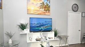A living room with a TV on a white shelf, a glass table, and a door. - Cute as a Button, AC, Starlink, Wi-Fi, Hot Tub, Near Beaches, Bars & Restaurants (North Palmetto Point)