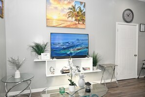 A living room with a TV on a white shelf, a glass table, and a door. - Cute as a Button, AC, Starlink, Wi-Fi, Hot Tub, Near Beaches, Bars & Restaurants (North Palmetto Point)