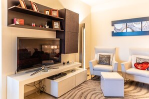 Ein modernes Wohnzimmer mit Flachbildfernseher, zwei weißen Sesseln, einem Couchtisch, einem Bücherregal und einem Gemälde an der Wand.