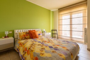 A bedroom with a bed, green walls, a window with blinds, and a nightstand with a lamp.