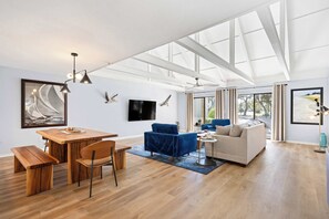 A spacious living area with a wooden dining table, blue armchairs, a sofa, and a large window providing natural light. - Fully Renovated Villa - Sleeps 6 Near Beach & Golf (Hilton Head Island)