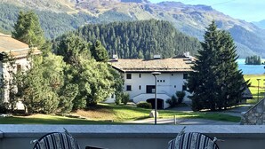 Una terraza con una computadora portátil, una botella y una taza sobre una mesa, rodeada de sillas a rayas, con vistas a una montaña y un pueblo.