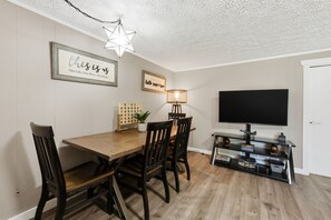 Un coin repas avec une table en bois, quatre chaises, un luminaire en forme d’étoile, une citation encadrée, une lampe et un téléviseur à écran plat posé sur un meuble.