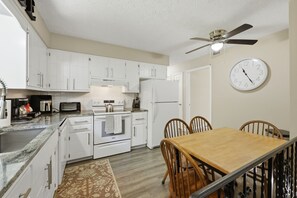 Une cuisine compacte avec des meubles blancs, une table à manger, des chaises, une horloge et un ventilateur de plafond.