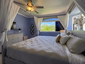 Quarto com cama, ventilador de teto, mesa de cabeceira, janela com cortinas e vista para o verde lá fora.