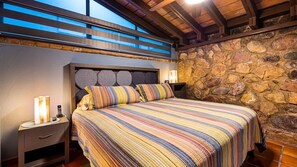 A bedroom with a stone wall, a bed with striped bedding, a nightstand with a lamp, and a wooden ceiling. - Zona Romantica Oasis/Pool/2 block Pier bars shop (Puerto Vallarta)