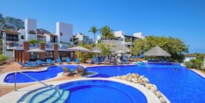 Una piscina amb gandules i ombrelles, envoltada d'edificis amb balcons i teulades de palla.