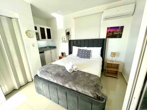 Ein Schlafzimmer mit einem Bett, einem Nachttisch, einer Uhr, einem Fenster und Blick auf eine Stadtansicht.