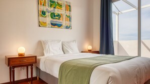 A hotel room with a bed, two bedside tables, a wall-mounted abstract painting, a window with a view, and a green throw blanket. - Verde Água Garajau, a Home in Madeira (Santa Cruz)