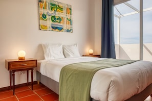 A hotel room with a bed, two bedside tables, a wall-mounted abstract painting, a window with a view, and a green throw blanket. - Verde Água Garajau, a Home in Madeira (Santa Cruz)