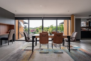 A modern dining area with a large table, six chairs, and a view of greenery through large windows.
